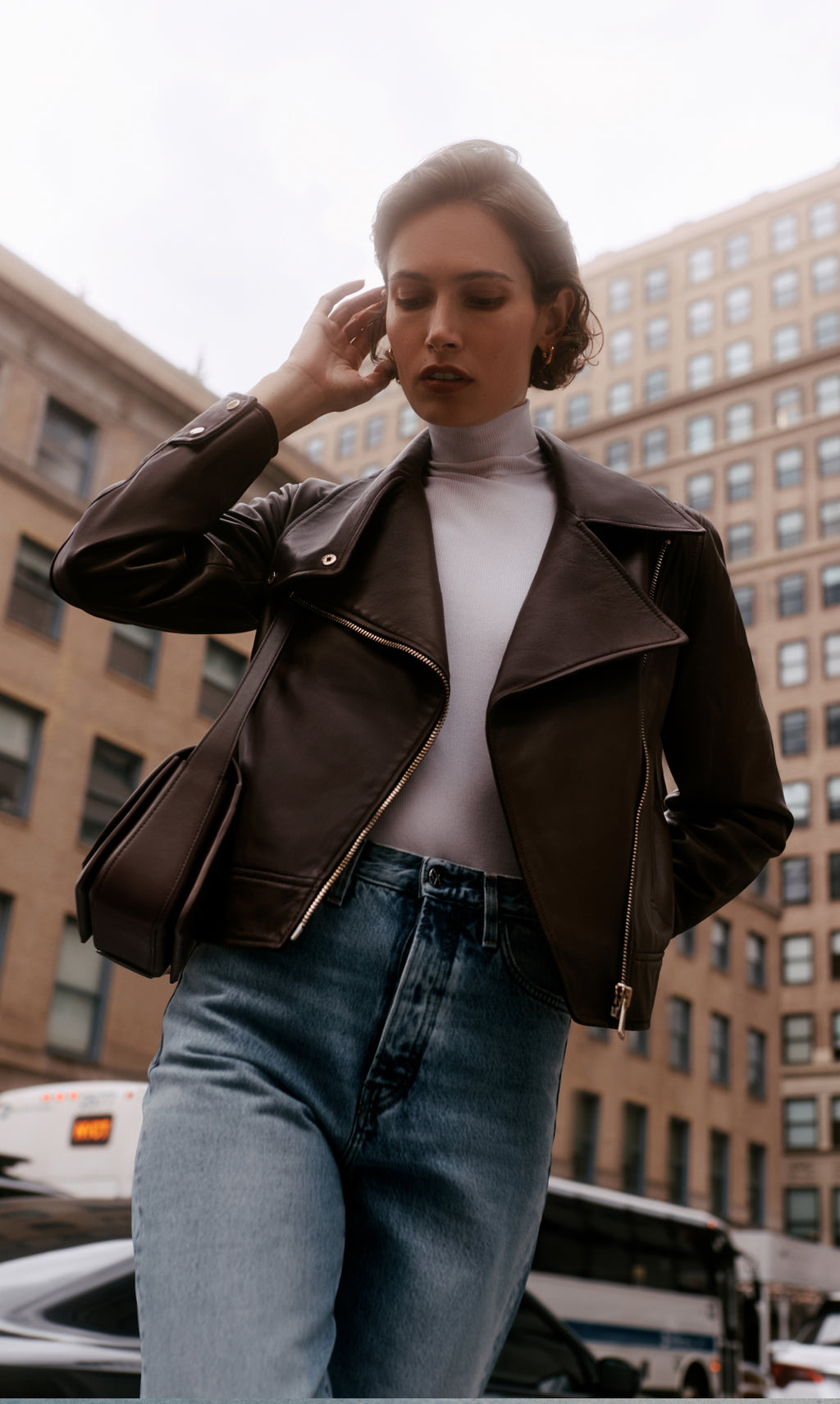 Person wearing a shoulder bag in a leather jacket and jeans stands on a city street with buildings in the background.
