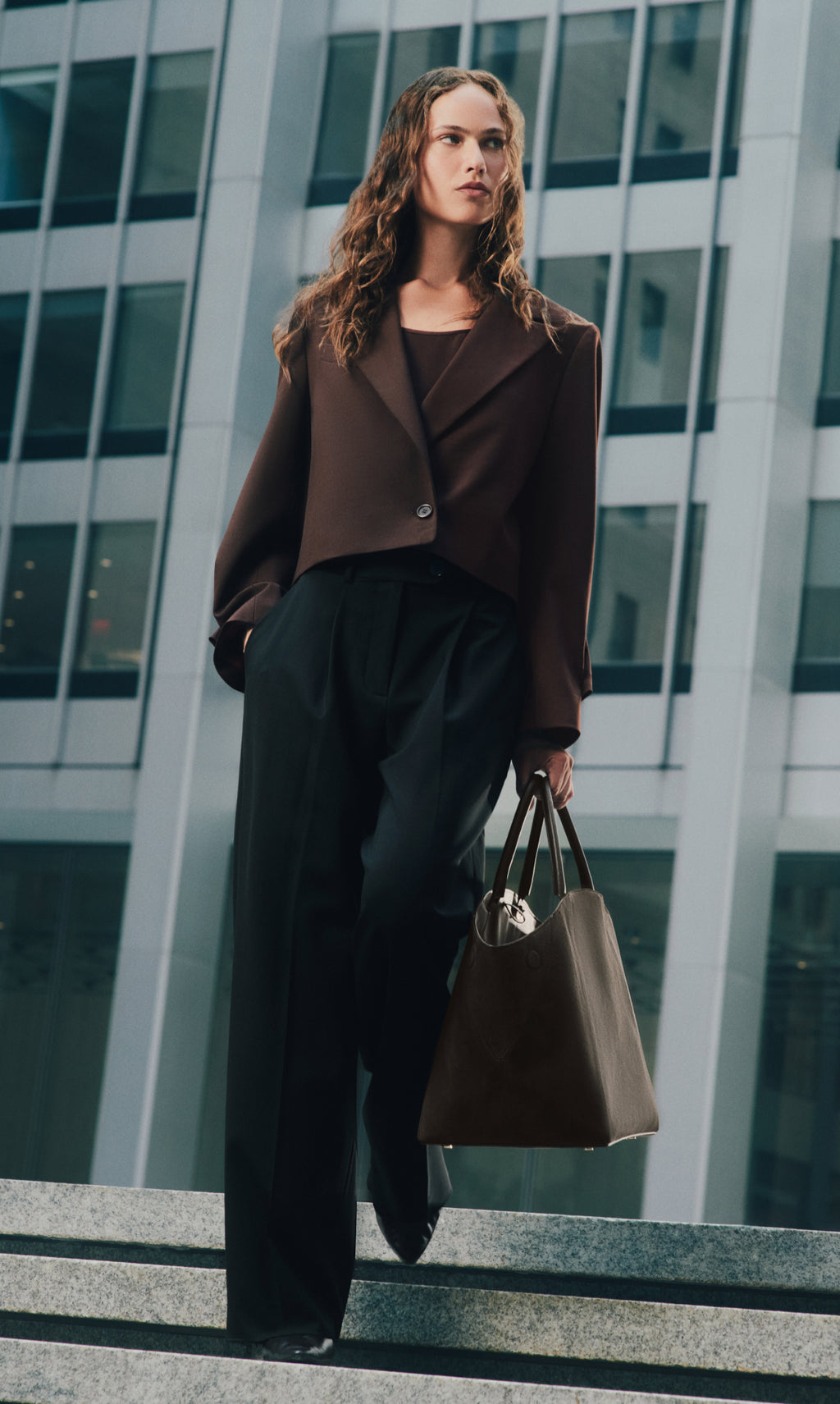 Person in a cropped jacket holding a large bag, standing near tall buildings.