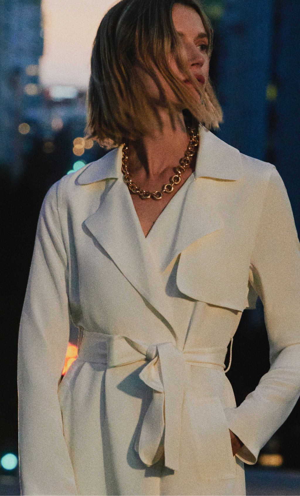 Person wearing a belted coat with a chunky chain necklace in an urban setting.