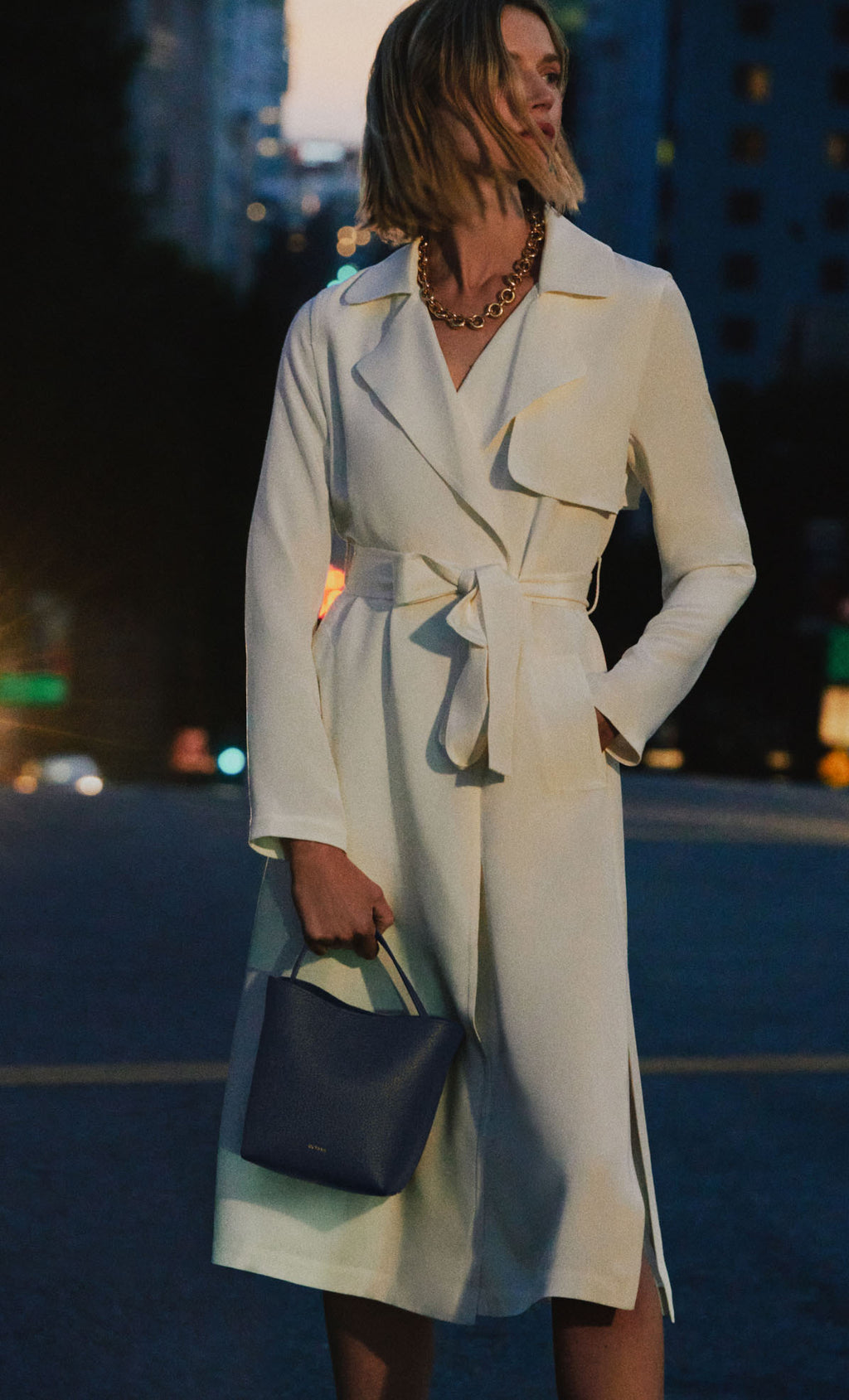 Woman in a belted coat holding a handbag on a city street.