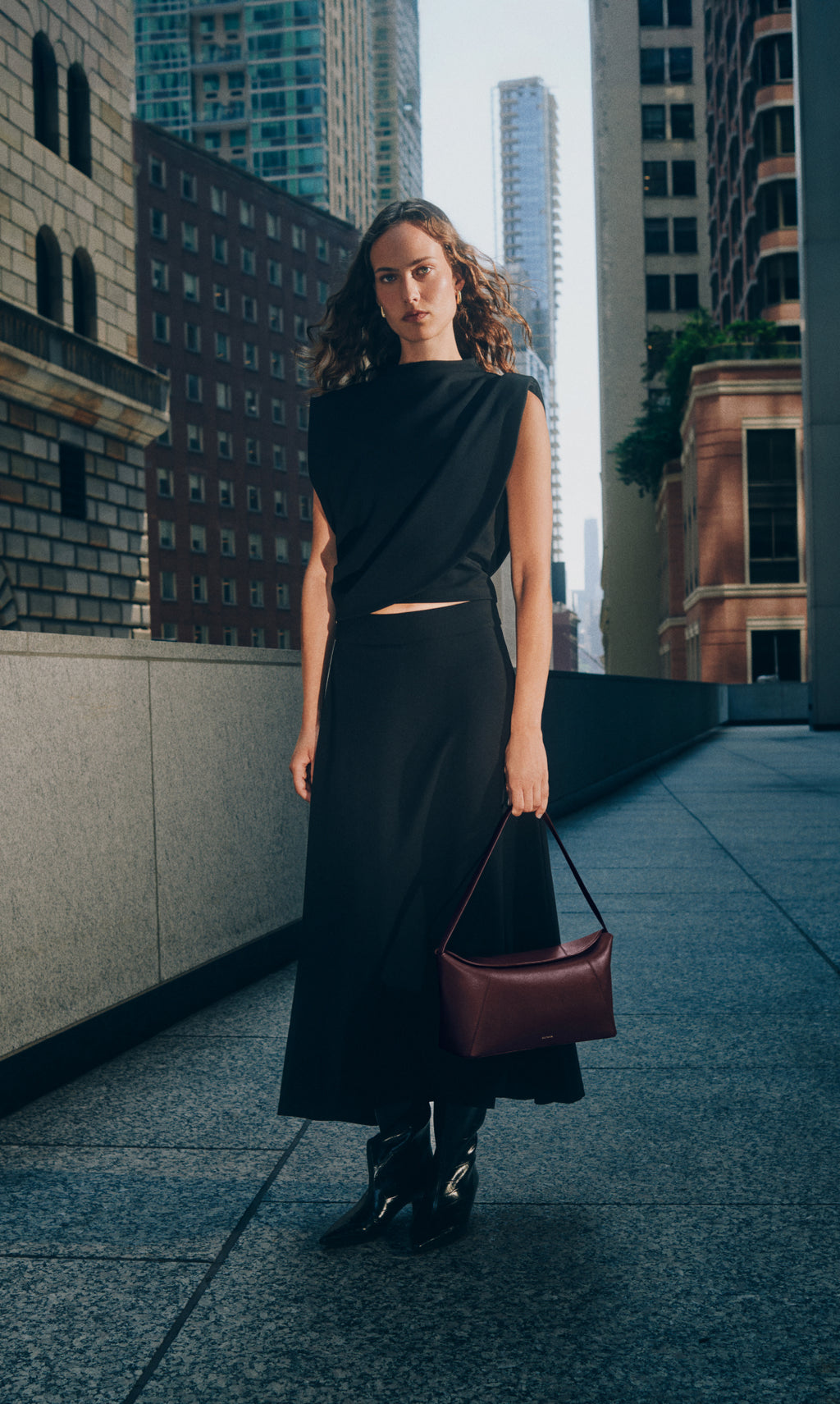 Person walking in city holding handbag, wearing long skirt, asymmetrical top and boots.