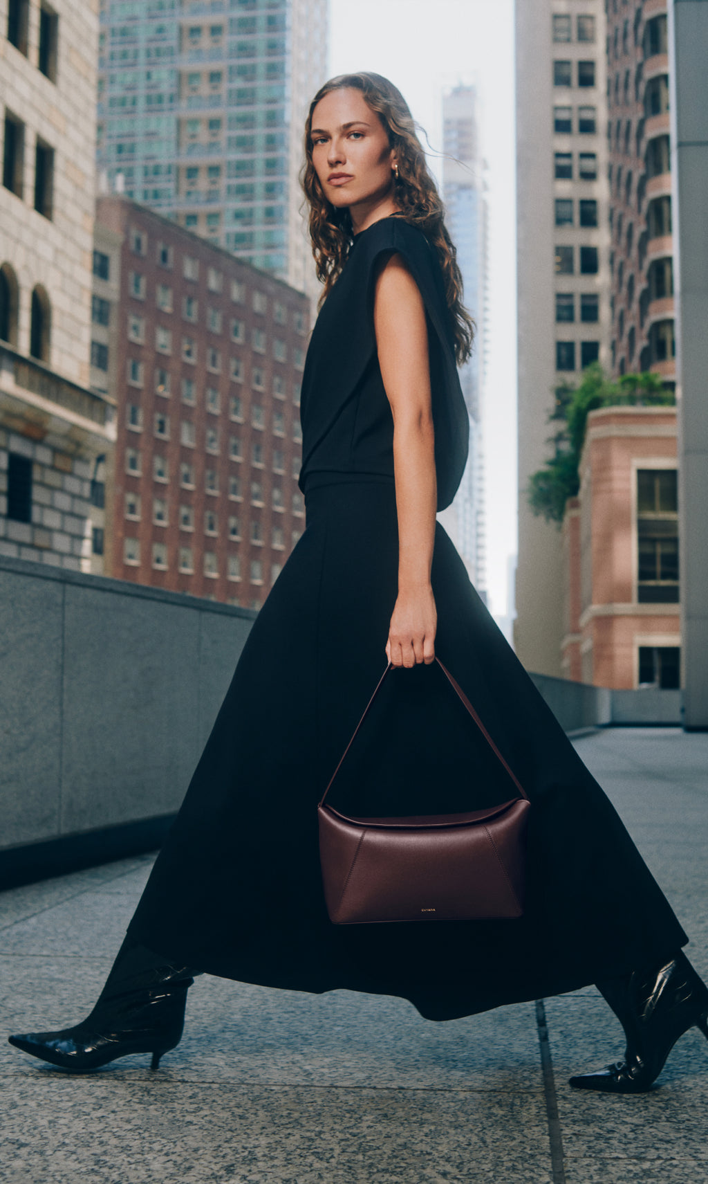 Person walking in city holding handbag, wearing long skirt, asymmetrical top and boots.