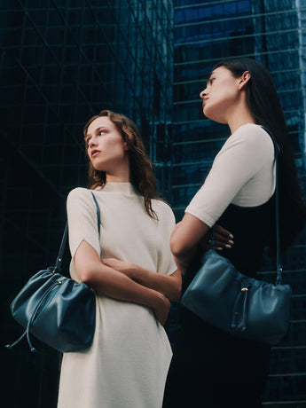Two people with shoulder bags stand outside tall glass buildings.