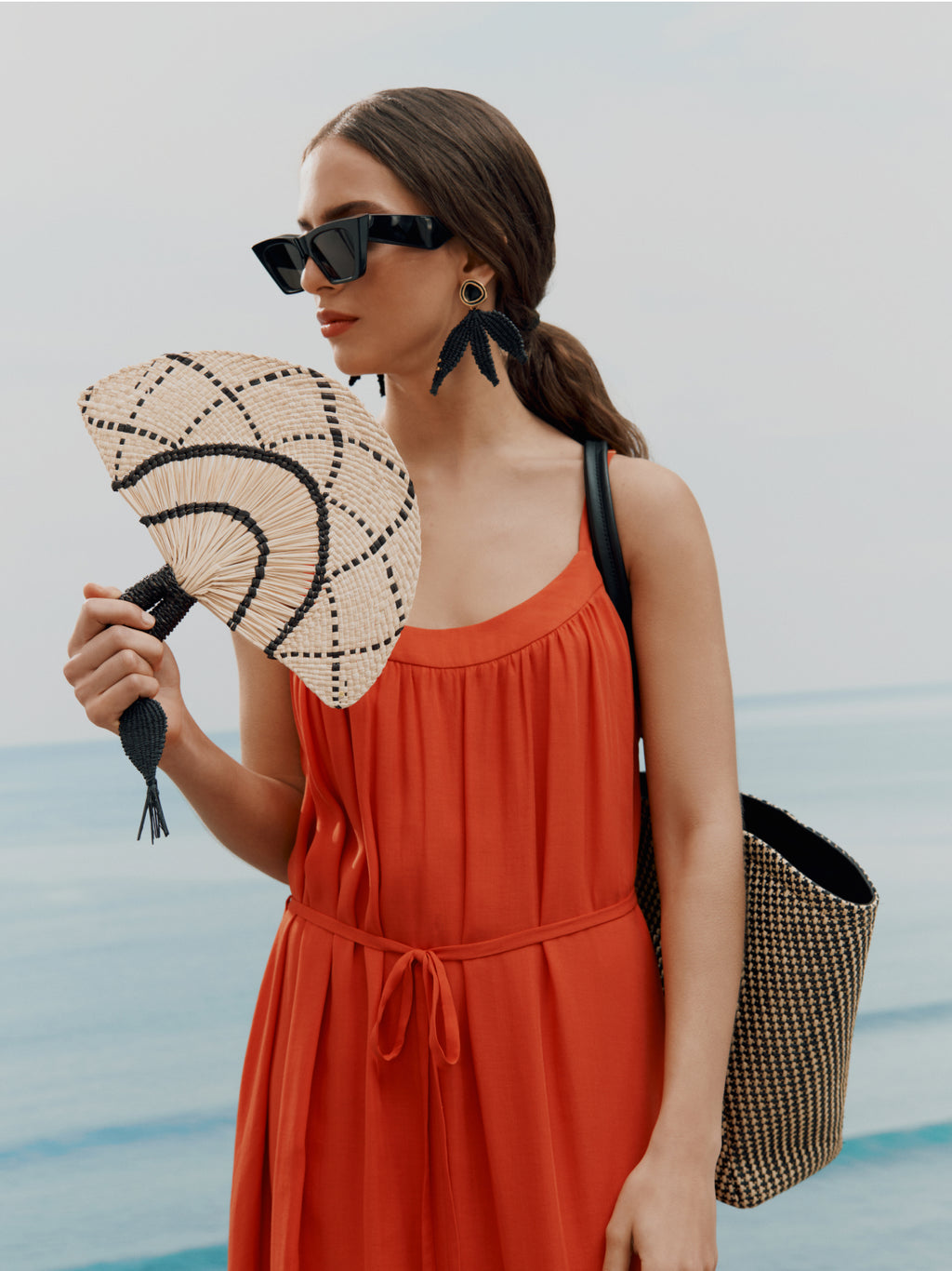 Woman in dress with fan, large earrings, sunglasses, and beach bag standing by the sea.