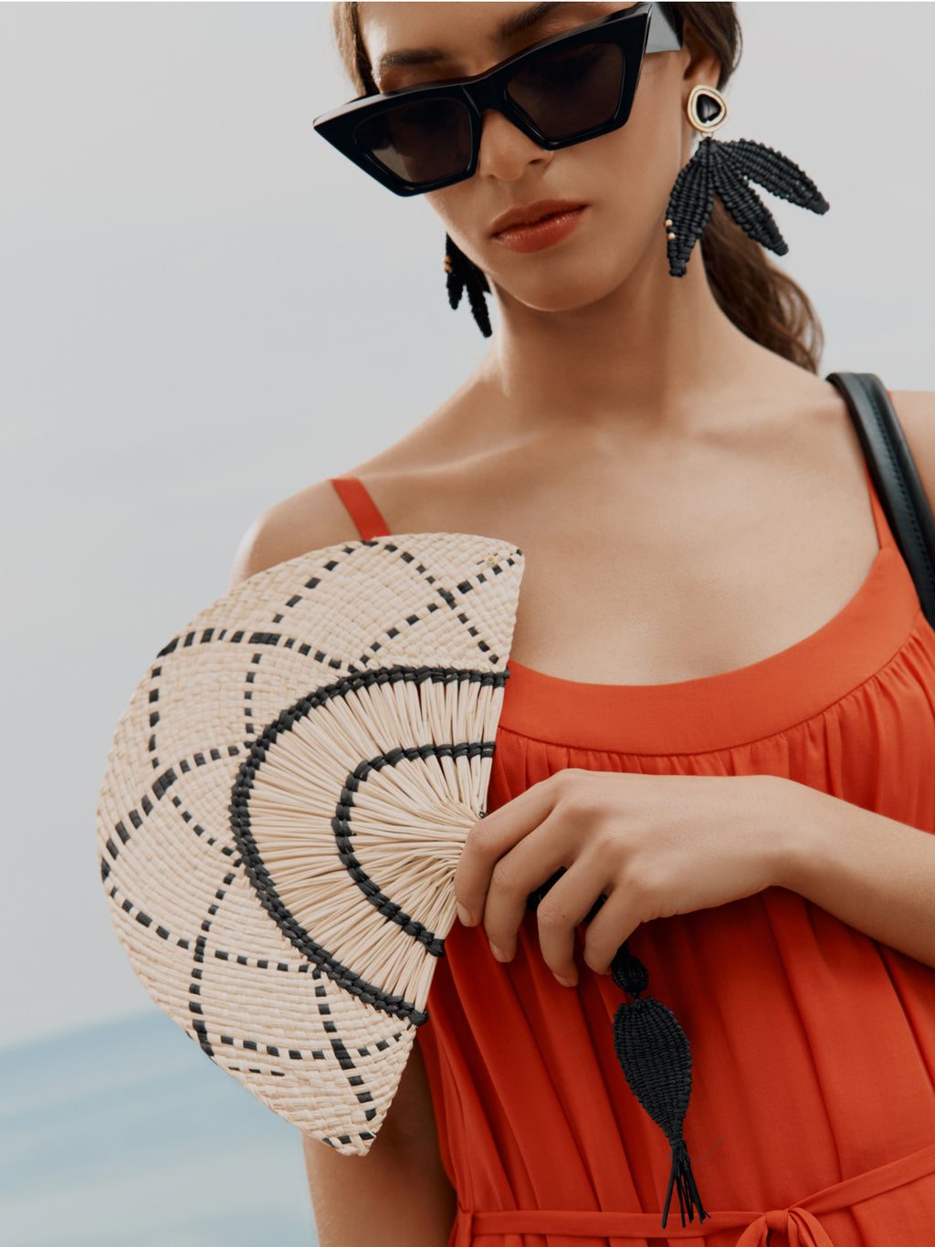 Woman holding a hand fan, wearing large sunglasses and earrings, with a relaxed outfit.
