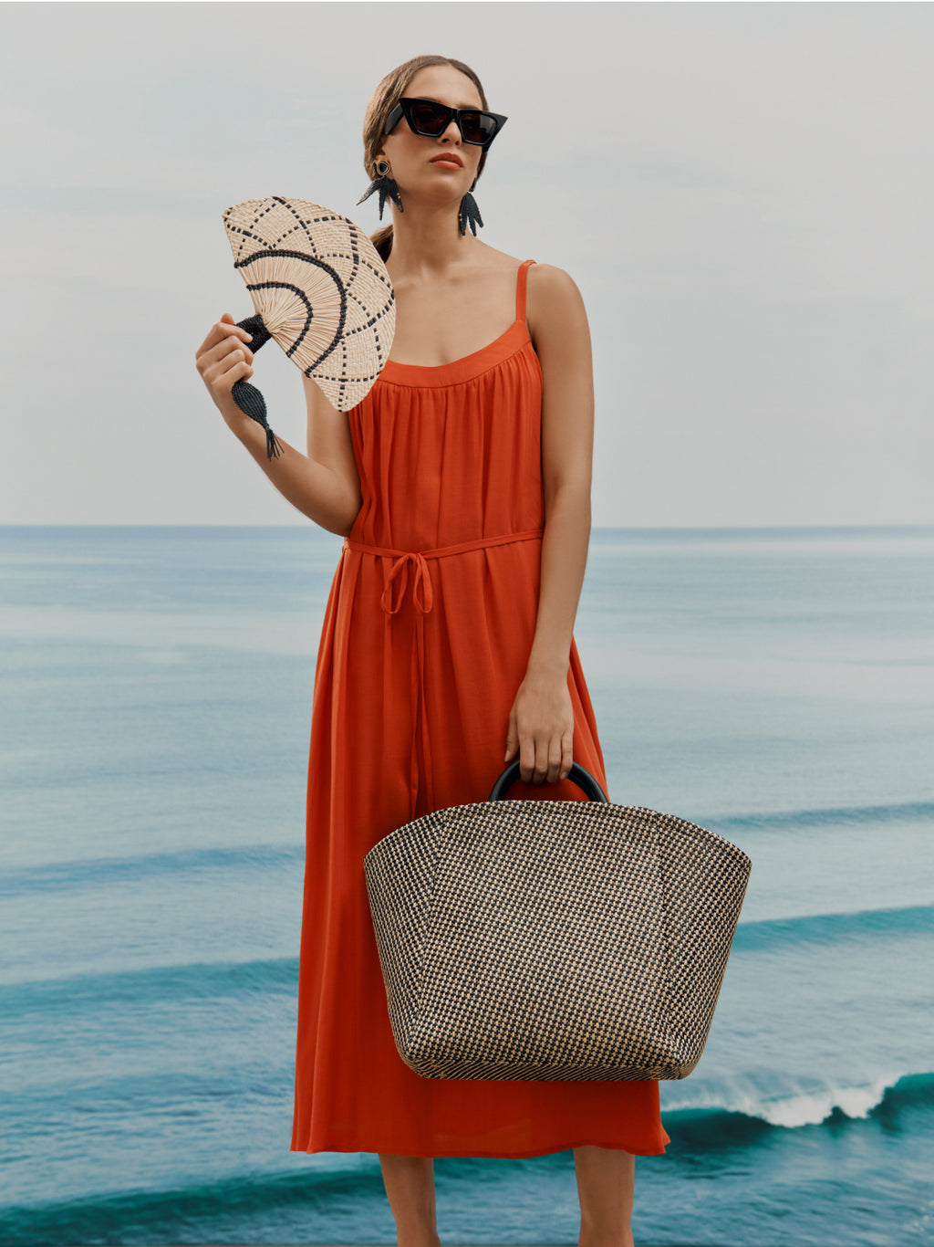 Person in a dress holding a hat and a large tote bag, standing in front of the ocean.
