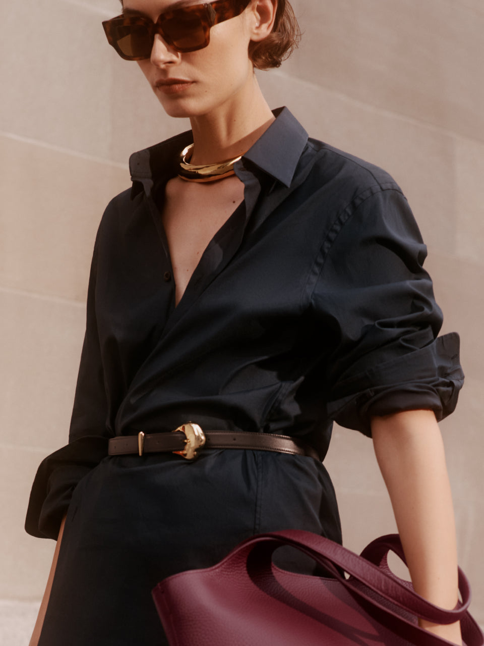 Person in a button-up shirt with rolled sleeves, wearing sunglasses and holding a purse.