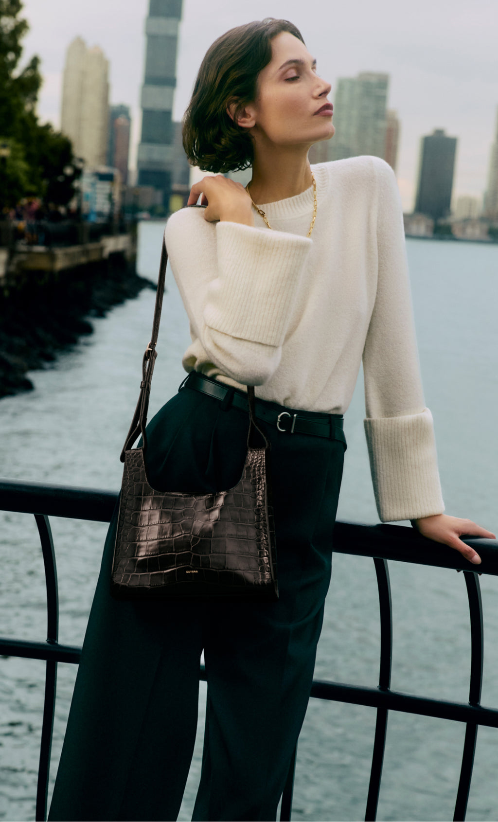 Woman leaning on railing, wearing sweater, pants, and carrying a croc-embossed leather handbag. Cityscape background.