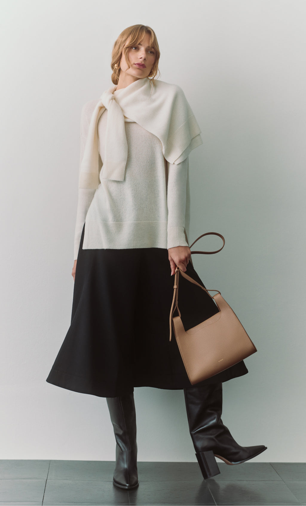 Person in a sweater and skirt holding a handbag, with boots on.