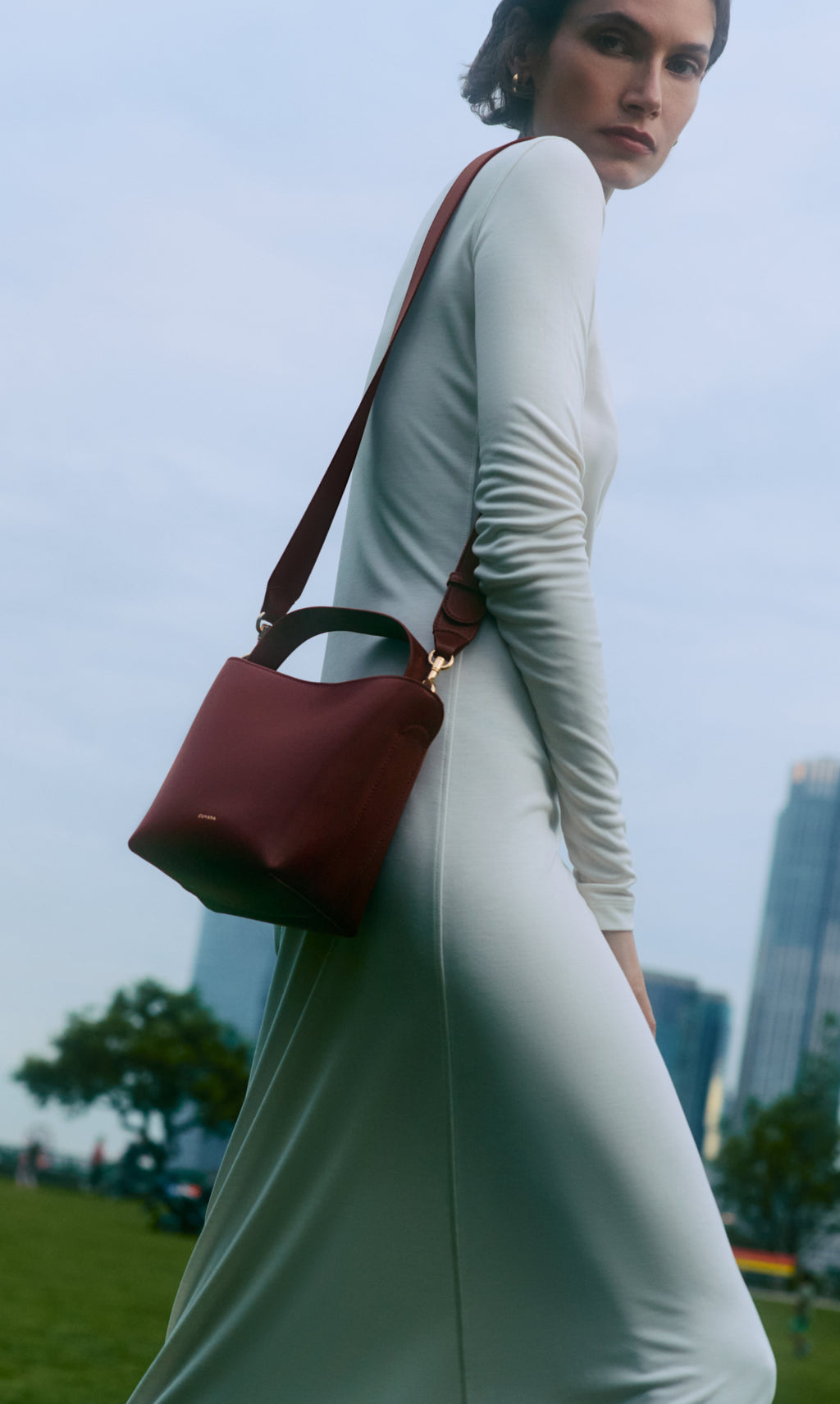 Person in a long dress carrying a shoulder bag, standing outdoors with buildings in the background