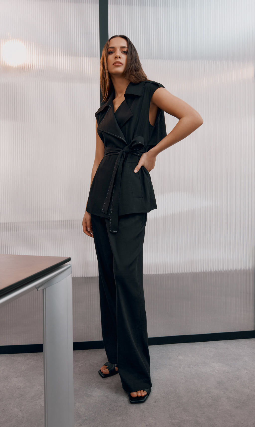 Woman in a sleeveless belted top and pants stands by a table indoors.