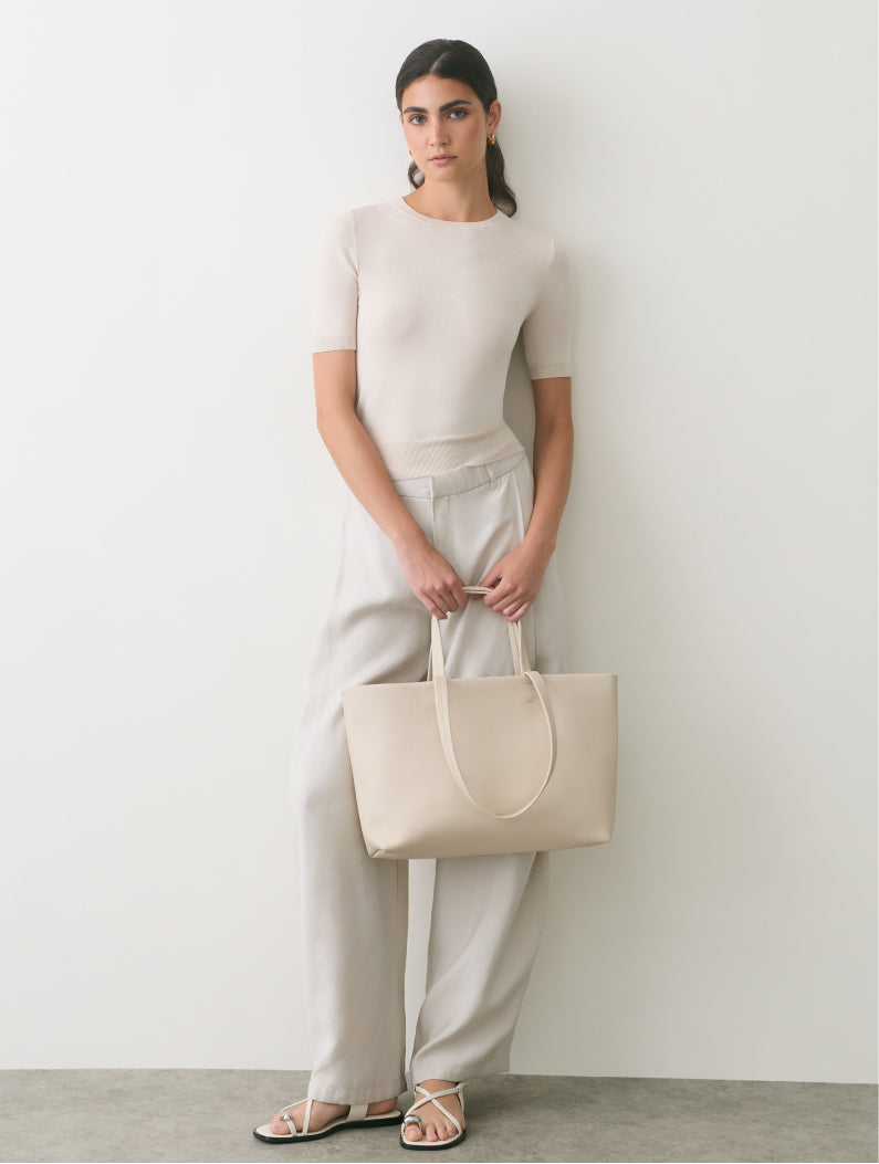 Person standing, holding a tote bag, wearing a top, pants, and sandals.
