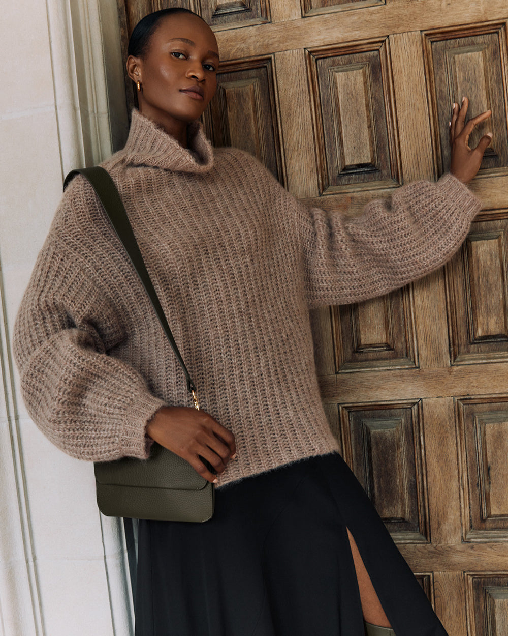 Person in a sweater and skirt with a shoulder bag, leaning against a wooden door.