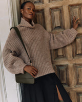 Person in a sweater and skirt with a shoulder bag, leaning against a wooden door.