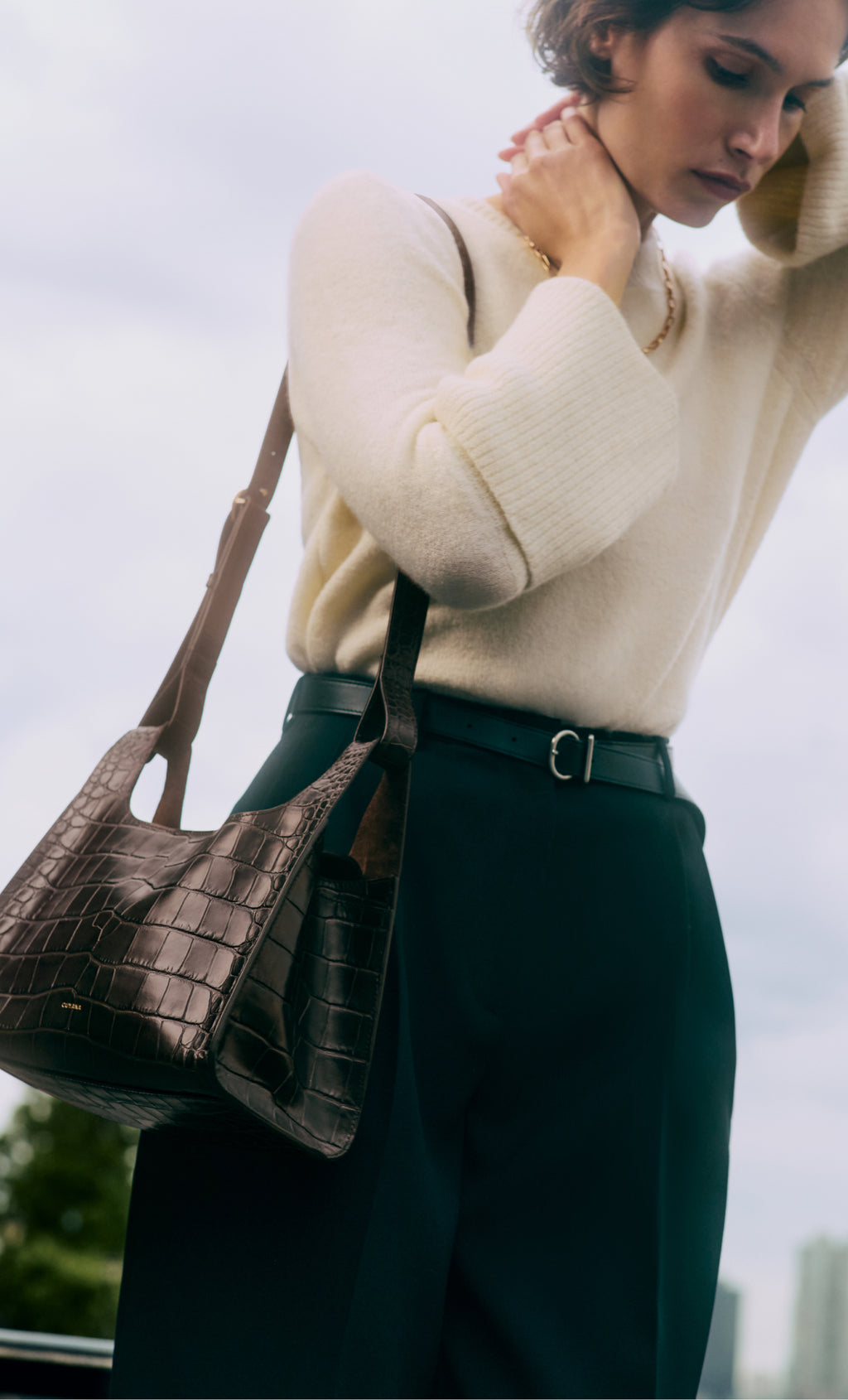 Person wearing sweater with croc-embossed leather shoulder bag and belted pants.