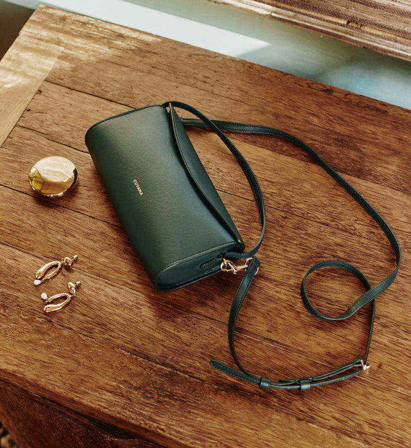 Handbag with a strap, near earrings and a round compact on a wooden table.