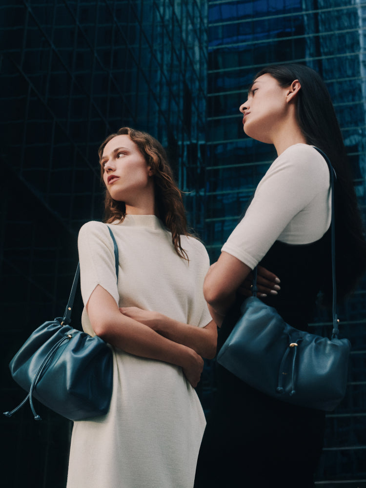 Two people each with a shoulder bag standing in front of tall buildings
