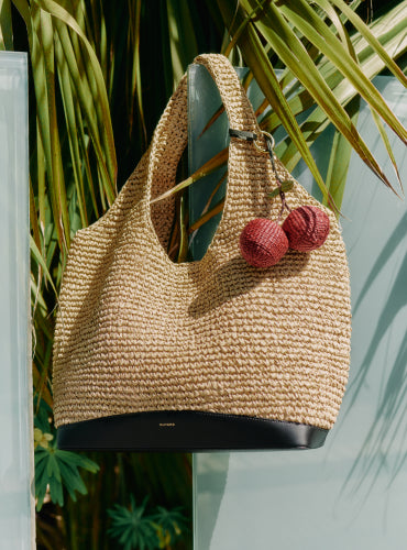 Woven handbag with pom-pom keychain hanging by plants.