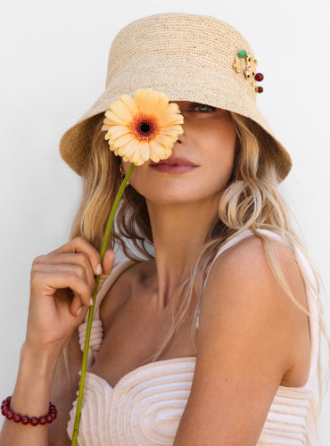 Woman wearing a hat holds a flower to her face, one eye visible.