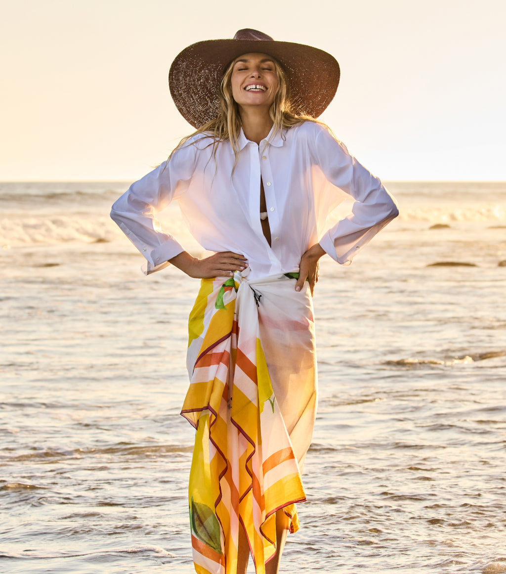 Person smiling at the beach wearing a wide-brimmed hat and holding a colorful wrap.