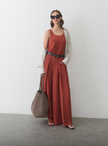 Woman in long dress with open shirt, sunglasses, holding a bag.