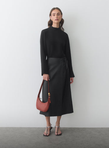 Person in long-sleeve top and skirt holding a shoulder bag, standing indoors.