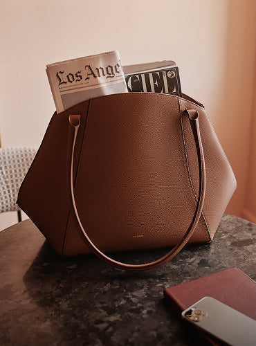 A tote bag with a newspaper and magazine on a table near a phone and a notebook.