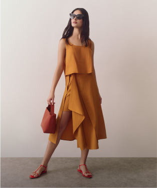 Woman in sleeveless dress holding handbag, wearing sandals and sunglasses.