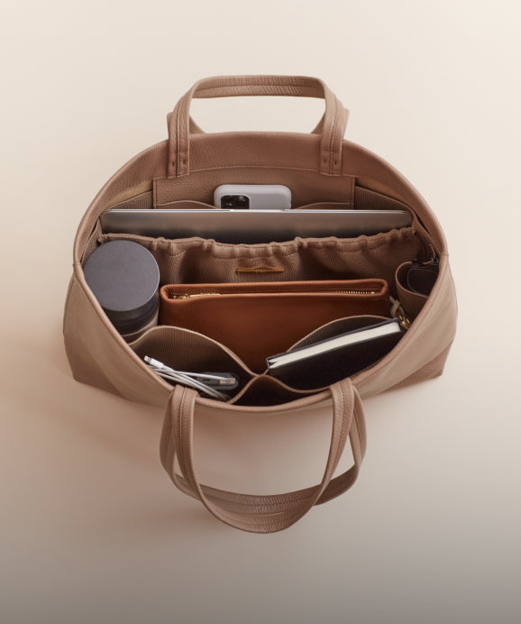 Linked Image: Directs to Bag Organizers shop page. Image Description: Interior of the Cuyana Easy Tote in Cappuccino with laptop, water bottle, notebook, phone charger and a Cuyana Zipper Pouch in color Caramel place inside. 