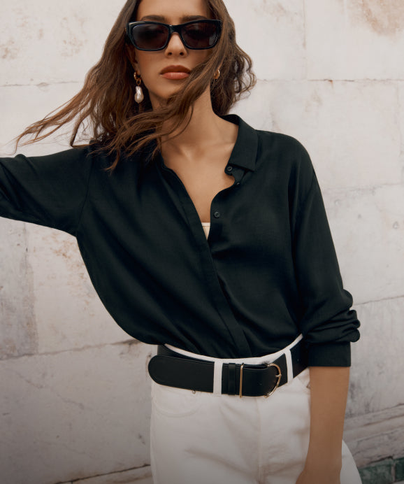 Woman posing in a buttoned shirt and belted trousers against a wall.