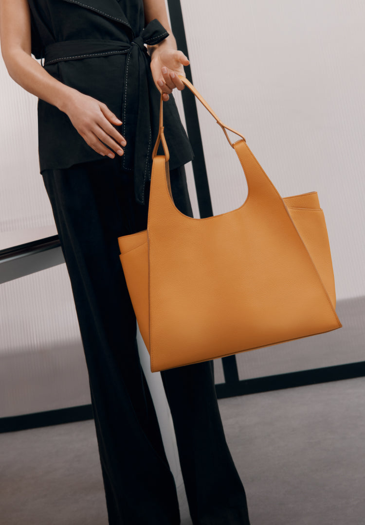 Model wearing black, holding the Cuyana Double Loop Satchel in color Mango. Button: 