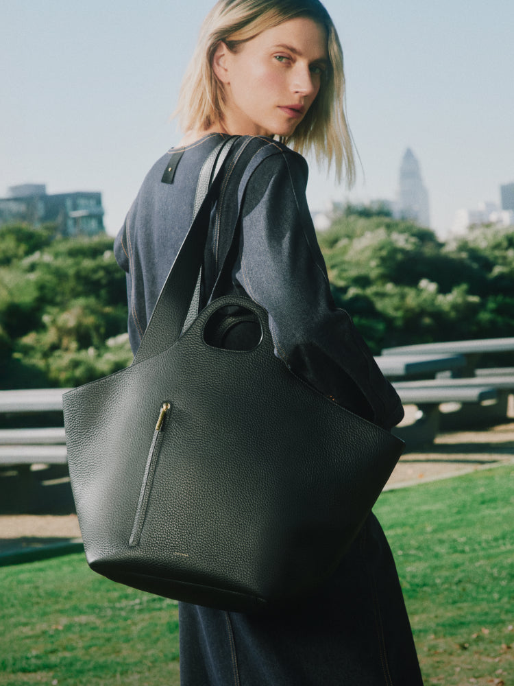 Person outdoors with large handbag over shoulder, city skyline in background.