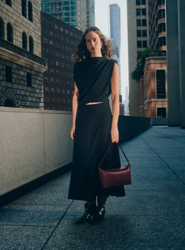 Person in sleeveless top and long skirt holding a handbag stands on an urban rooftop walkway.