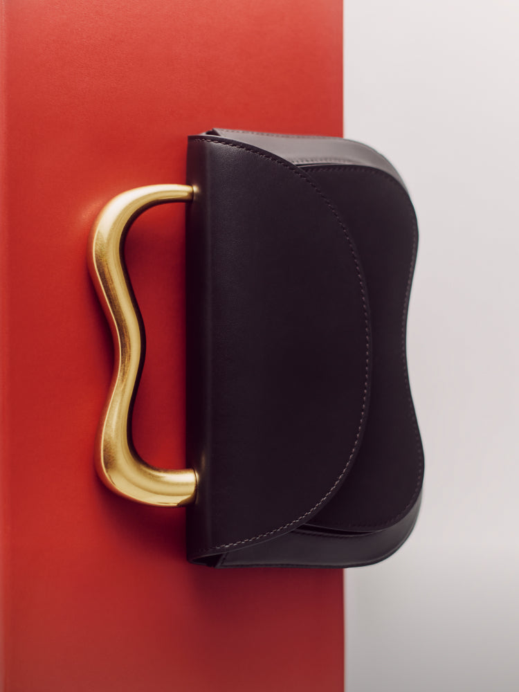 A smoother leather handbag with a wavy metal handle laying on a red surface. 