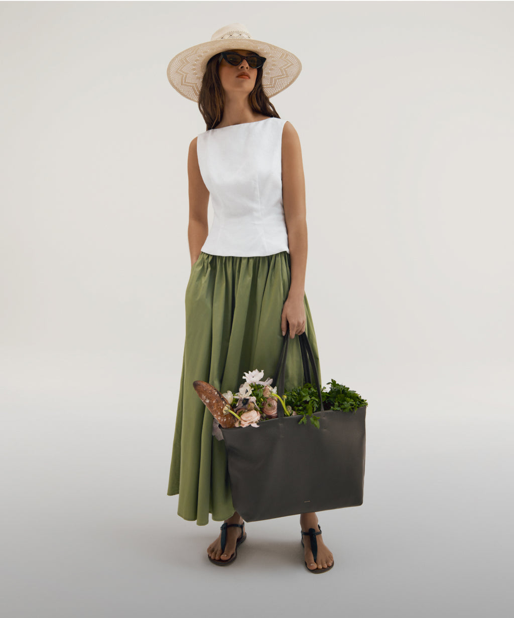 Linked Image: Directs to Summer Collection preview, Image Description: Model wearing Cuyana Open Weave Ecuador Hat in color Natural, Poplin Gathered Skirt in color Sage and a white top, carrying the Cuyana Easy Tote in color Dark Olive. 
