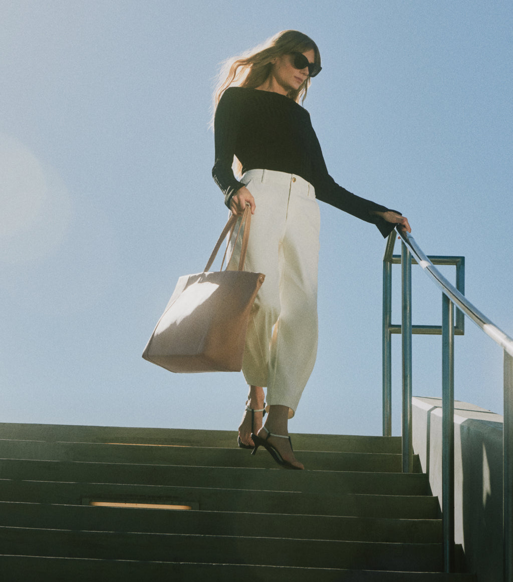 Person walking down steps holding a large tote bag, wearing sunglasses and heeled sandals.