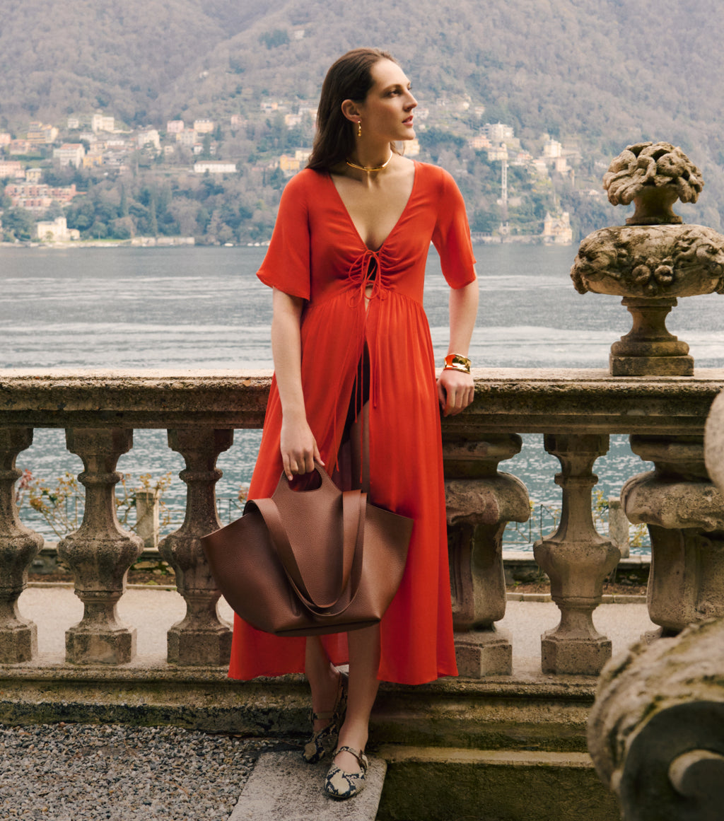 Person in dress stands by railing with large bag and scenic view behind.
