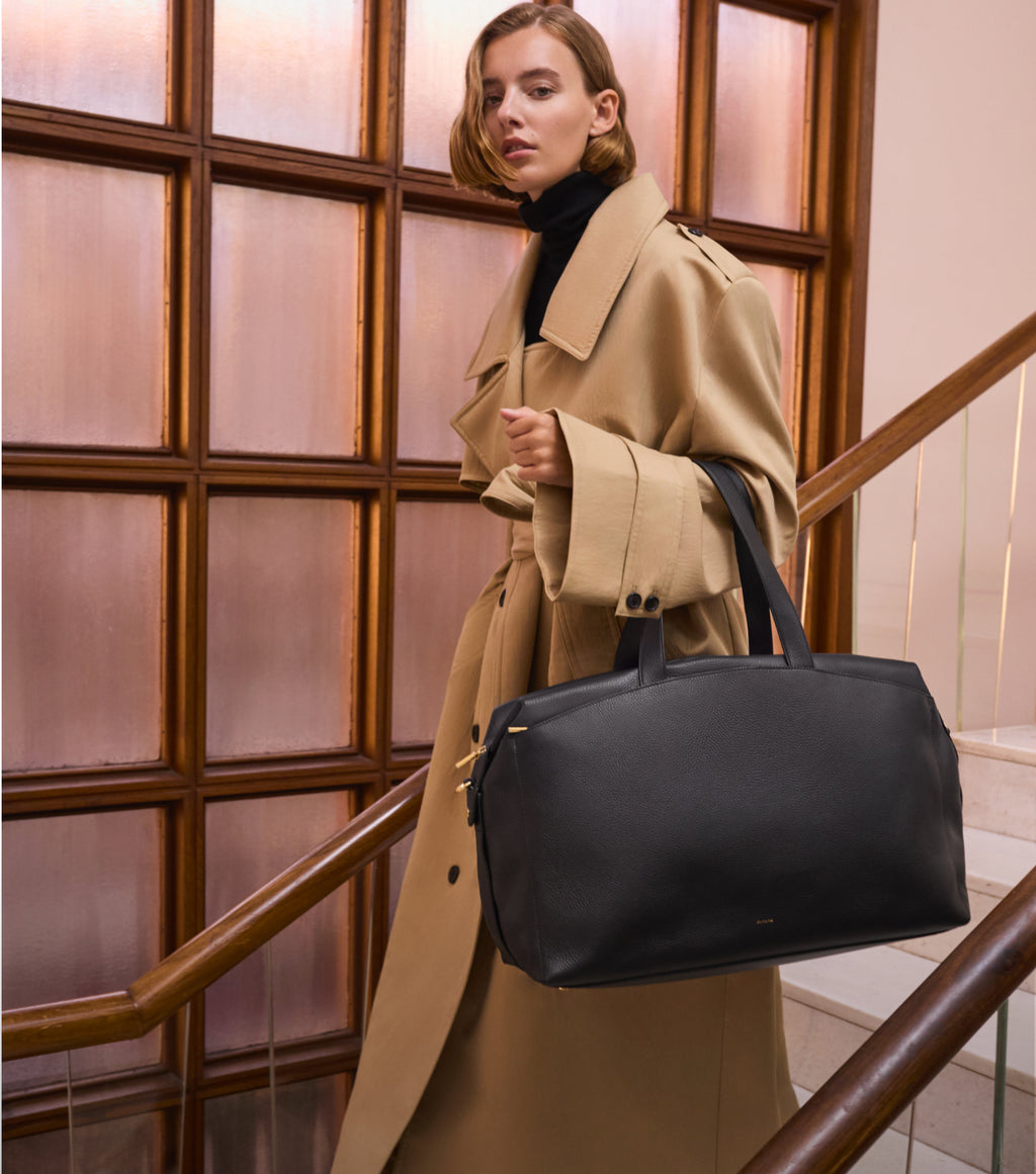 Person wearing a trench coat and carrying a large travel bag walks down stairs. 