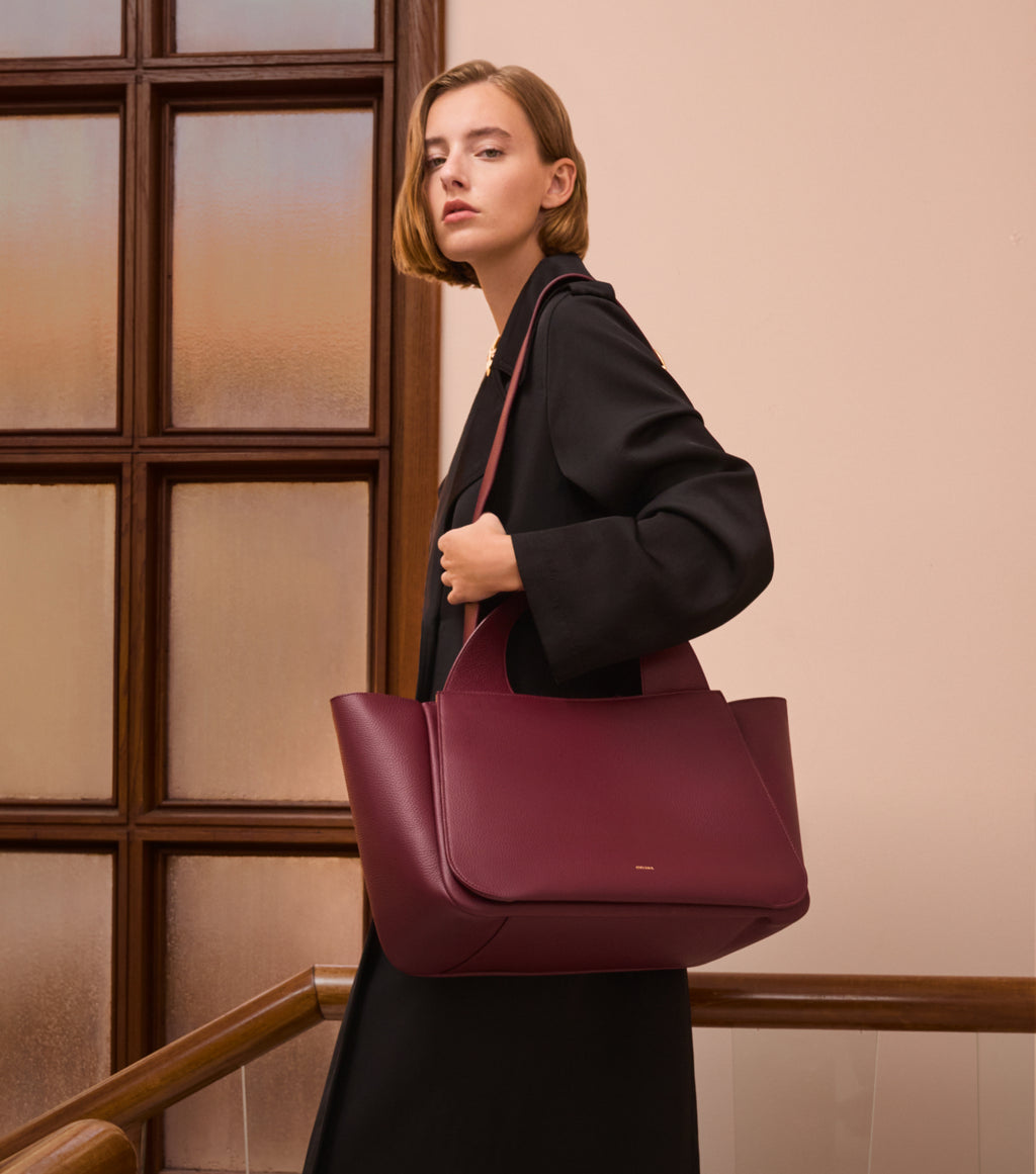 Person carrying large handbag, standing indoors by a window.