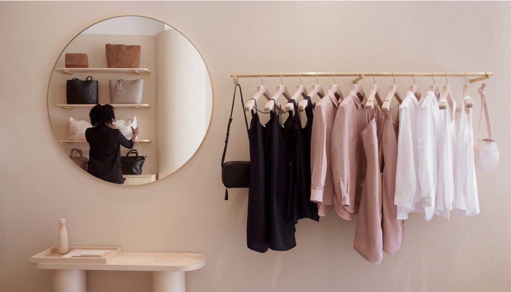 Linked Image: Image directs to information on retail locations. Image Description: Retail wall showing a leather handbag in black and clothing in black, pink and white. A mirror reflects a person wearing black and leather totes in black, taupe and brown. 