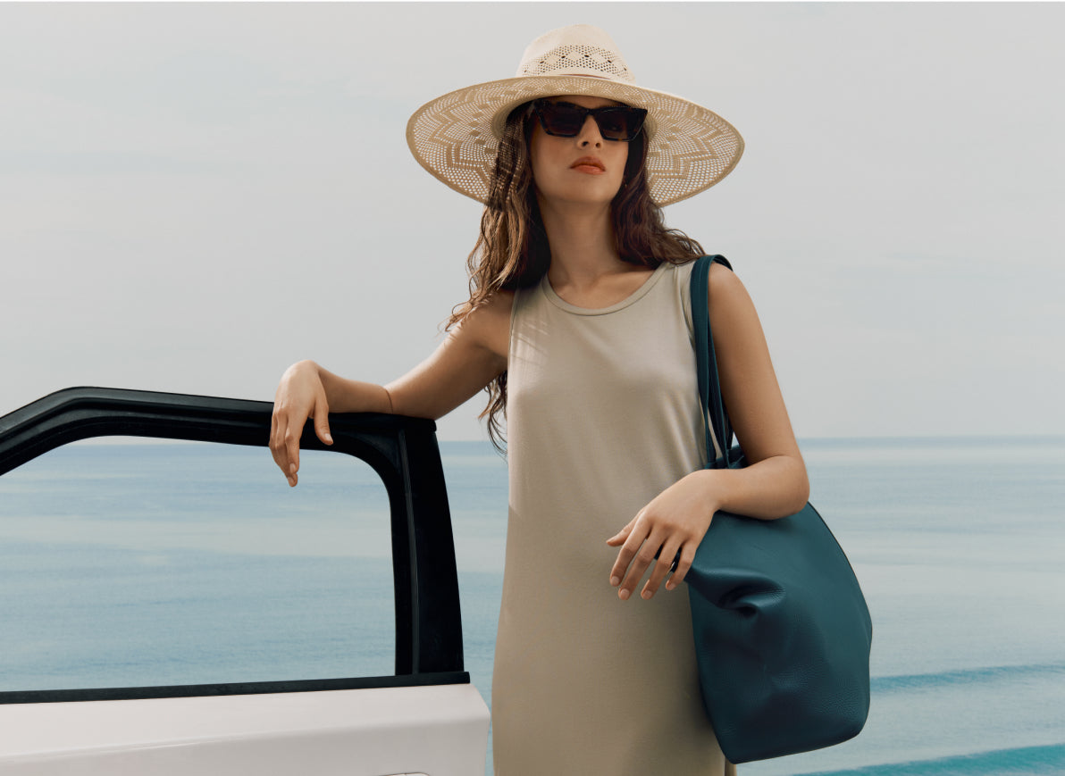 Woman in a hat and sunglasses standing by a car with the ocean in the background.