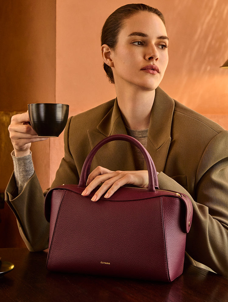 Person seated at a table holding a cup and a handbag.