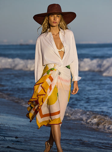 Person in a white shirt and patterned skirt walks on the beach wearing a wide-brimmed hat.