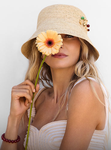Person wearing a hat holding a flower in front of one eye.