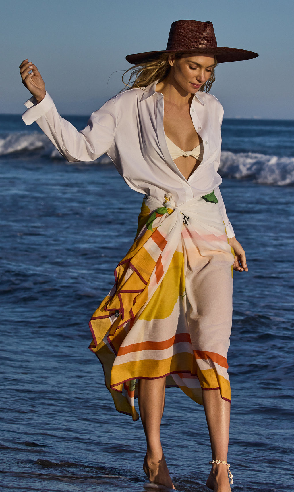 Person walking on beach wearing hat, open shirt, and striped sarong.