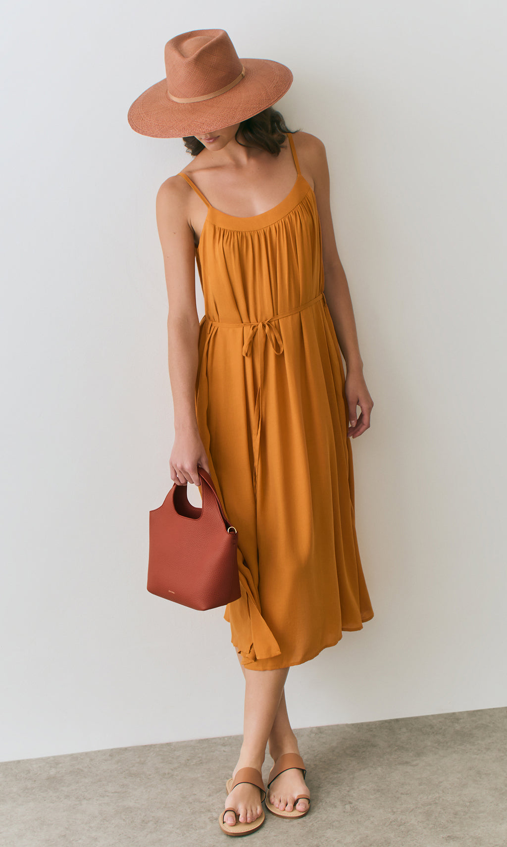 Model wearing a long dress and a wide-brim hat, holding a small handbag.