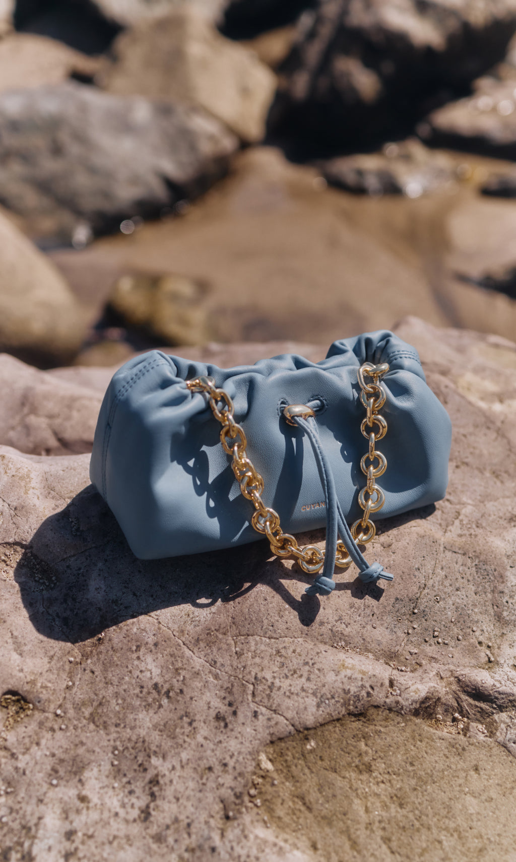 Handbag with chain strap on rocks near water.