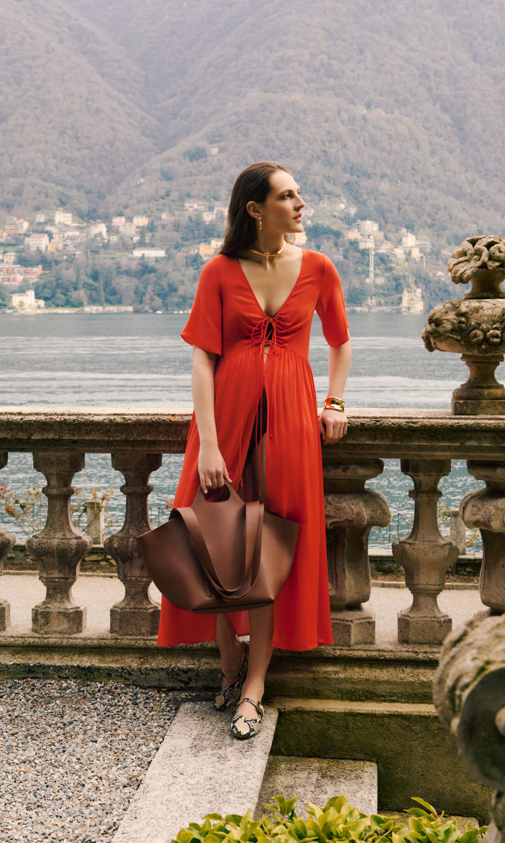Woman standing by stone railing with lake and mountains in background, holding large bag
