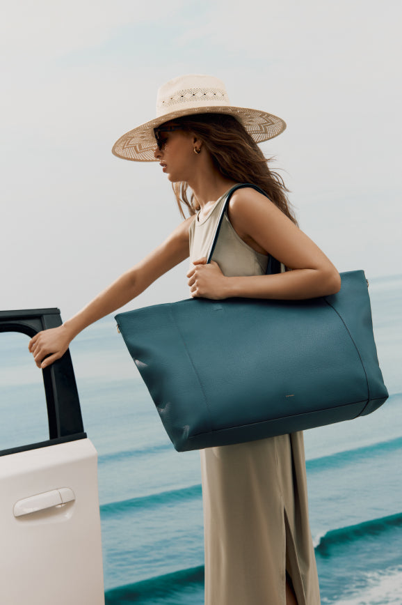 Woman holding large tote bag, standing next to an open car door, ocean in the background