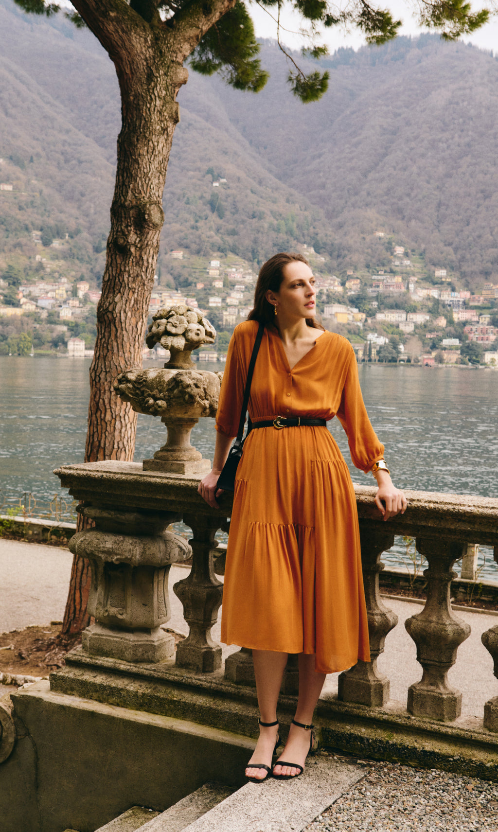 Woman in dress stands by stone railing overlooking a lake and mountains